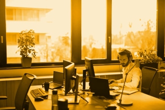 Man wearing headphones working at a computer desk in a modern office with large windows and plants.